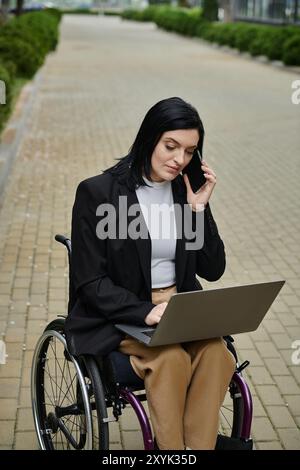 Una donna su una sedia a rotelle lavora sul suo computer portatile mentre parla al telefono. Foto Stock