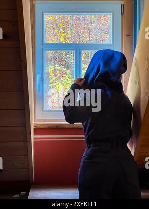 Una donna con una calda felpa con cappuccio blu si erge e guarda fuori dalla finestra in autunno in un'accogliente cabina di legno, in una tranquilla mattinata d'autunno, determinando il W Foto Stock