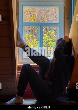 Una donna con una calda felpa con cappuccio blu si siede sul davanzale e guarda fuori dalla finestra in autunno in un'accogliente cabina di legno, in una tranquilla mattinata d'autunno, d Foto Stock