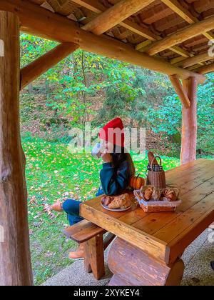 Una donna dai lunghi capelli scuri, con un berretto rosso brillante e una tuta verde, si siede su una panchina vicino al tavolo all'esterno di una rustica cabina in legno. L'atmosfera Foto Stock