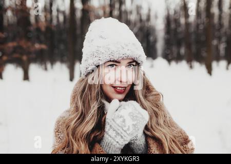 Bella giovane donna in neve fantasia bosco invernale. Ragazza che indossa morbidi guanti, cappuccio e cappotto. Foresta di Natale, alberi su sfondo sfocato. Incrociato Foto Stock