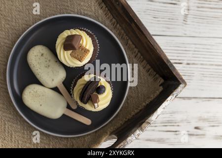 Biscotti bianchi, gelati su un bastone e gustosi muffin e cupcake su un vassoio su sfondo di legno. Decorate con caramelle, biscotti e colo diversi Foto Stock