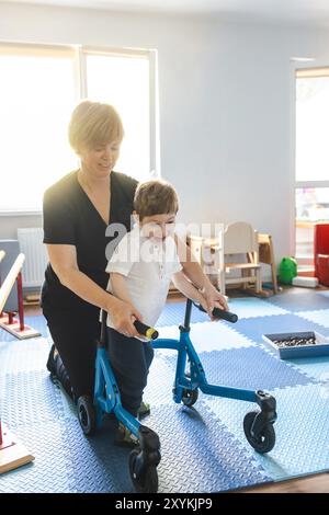 Il fisioterapista assiste un bambino con disturbi del movimento mentre si impegna nella riabilitazione fisica utilizzando un deambulatore in una sala di terapia luminosa Foto Stock