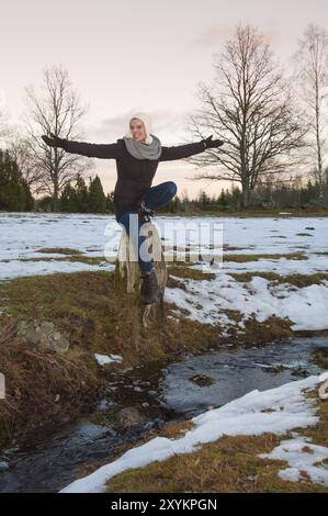 Giovane donna seduta su un ceppo d'albero vicino a un piccolo torrente e godendo il tardo inverno. Lo scioglimento della neve annuncia la fine dell'inverno Foto Stock
