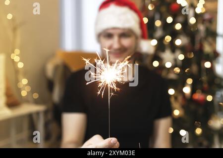 Il giovane uomo in Santa Hat sta tenendo scintillio ardente vicino all'albero di Natale. Luci di Natale in mani di ragazzo su sfondo sfocato con decorazioni di Natale t. Foto Stock