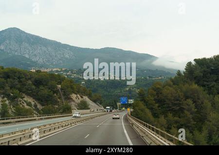 Mentone, Francia - 6 giugno 2023: Un'auto guida lungo un'autostrada, passando attraverso una regione montana panoramica con cime nebbiose e valli verdeggianti. Foto Stock