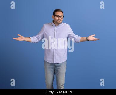 Un uomo d'affari confuso che allarga le mani e le spalle mentre si trova su sfondo blu Foto Stock