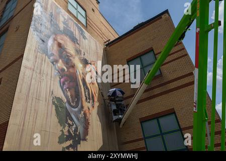 Southend on Sea, Regno Unito. 30 agosto 2024. Un grande murale viene dipinto sul lato di un edificio in mattoni, caratterizzato da un ritratto vibrante ed espressivo. Un artista è sollevato su un sollevatore idraulico verde, lavorando attivamente sulle sezioni superiori dell'opera. L'edificio presenta mattoni color marrone e rosso con finestre incorniciate di verde, che catturano il processo dinamico di creazione dell'arte pubblica. City Jam apre ufficialmente, con oltre 200 artisti provenienti da tutto il mondo che dipingono murales. Penelope Barritt/Alamy Live News Foto Stock