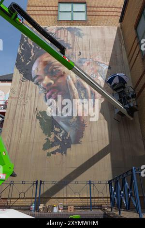 Southend on Sea, Regno Unito. 30 agosto 2024. Un grande murale che presenta un ritratto dettagliato viene dipinto sul lato di un edificio. L'artista lavora da un ascensore meccanico ombreggiato da un ombrello, con forniture di vernice visibili sotto. Il murale è parzialmente completo e mostra il processo di creazione della Street art. City Jam apre ufficialmente, con oltre 200 artisti provenienti da tutto il mondo che dipingono murales. Penelope Barritt/Alamy Live News Foto Stock