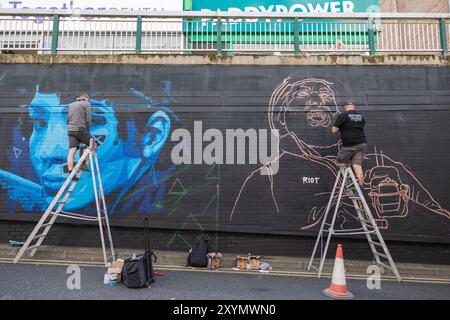 Southend on Sea, Regno Unito. 30 agosto 2024. Due artisti lavorano su un grande murale in corso su un muro di mattoni. Un lato presenta un ritratto di colore blu con ombreggiatura geometrica; l'altro mostra una figura disegnata. Scale, materiali di verniciatura e un cono segnalano l'area di lavoro attiva. La segnaletica sopraelevata include "PADDYPOWER". City Jam apre ufficialmente, con oltre 200 artisti provenienti da tutto il mondo che dipingono murales. Penelope Barritt/Alamy Live News Foto Stock