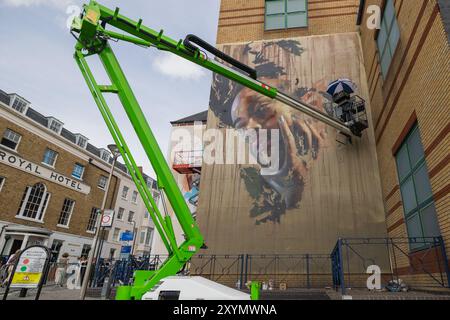 Southend on Sea, Regno Unito. 30 agosto 2024. Un vivace murale in corso sul lato di un edificio in mattoni a piu' piani a Southend-on-Sea. L'opera presenta un ritratto gioioso, con l'artista sollevato su un braccio articolato verde, che dipinge attivamente. La scena include recinzione temporanea, pedoni e il vicino Royal Hotel. City Jam apre ufficialmente, con oltre 200 artisti provenienti da tutto il mondo che dipingono murales. Penelope Barritt/Alamy Live News Foto Stock