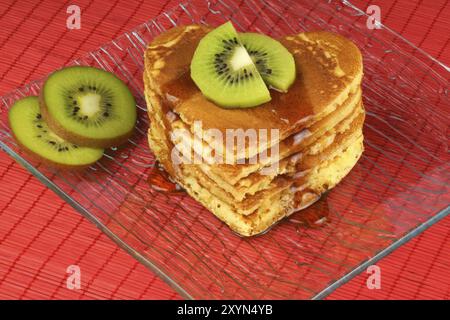 Pancake a forma di cuore con sciroppo e kiwi su un piatto di vetro trasparente su sfondo rosso. Una colazione perfetta per San Valentino Foto Stock