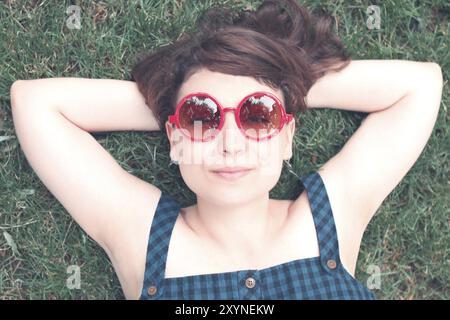 Dall'alto di una giovane donna felice e rilassata con occhiali da sole rotondi alla moda che guarda la macchina fotografica mentre sdraiata sull'erba verde e si gode la giornata estiva Foto Stock