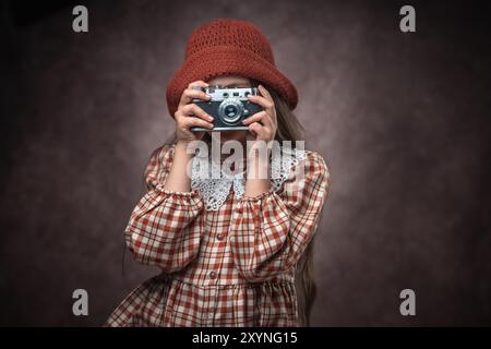 Ritratto retrò di una ragazza caucasica vestita con abito vintage e cappello con vecchia fotocamera retrò su sfondo marrone in pelle Foto Stock