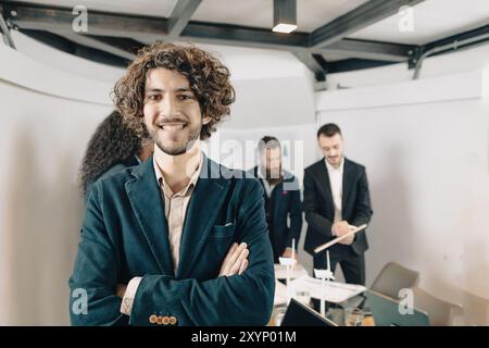 Il giovane architetto sorride con le braccia incrociate mentre il suo team sta lavorando sullo sfondo Foto Stock