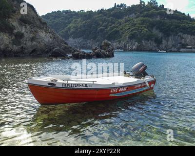 Una barca rossa galleggia in sicurezza nelle acque cristalline con strutture rocciose e verdi montagne intorno, corfù, mediterraneo, grecia Foto Stock