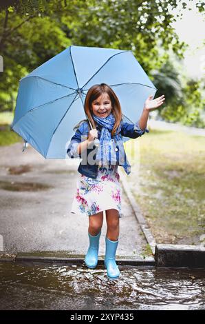 Giocoso eccitato bambino prescolare in abiti casual e stivali di gomma con ombrello blu ridere e saltare in pozza sorridendo alla macchina fotografica durante il gioco Foto Stock