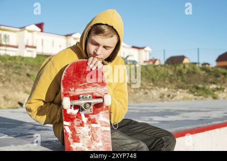 Primo piano di un giovane hipster vestito con una felpa e jeans seduti tristi in uno skate Park e con in mano uno skateboard Foto Stock