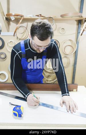 Un bel falegname stanco con barba su un tavolo con una matita disegna un cartello sul tabellone. Sul posto di lavoro c'è un normale maestro vestito di blu CA Foto Stock