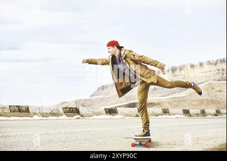 Elegante giovane felice con un berretto e pantaloni che jogger girovagano lungo una strada di montagna su un longboard, godendosi la vita Foto Stock