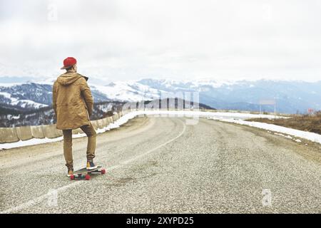 Elegante giovane felice con un berretto e pantaloni che jogger girovagano lungo una strada di montagna su un longboard, godendosi la vita Foto Stock