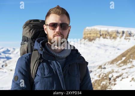 Un viaggiatore hipster con la barba che indossa occhiali da sole in natura. Un uomo che cammina in montagna con uno zaino e bastoni da passeggio scandinavi nel bac Foto Stock