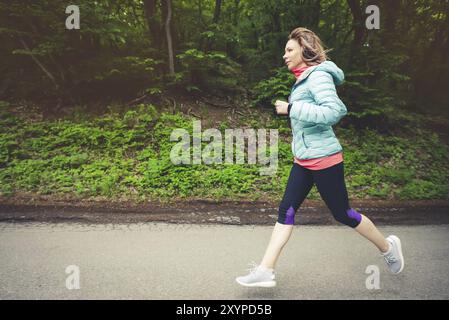 Una giovane donna bionda che corre si pratica all'aperto in un parco montano cittadino nella foresta. Raggi caldi attraverso i rami degli alberi Foto Stock