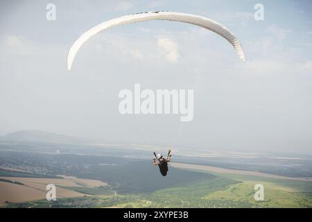 Il parapendio professionista in tuta di cocco vola in alto sopra il terreno contro il cielo e i campi Foto Stock