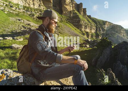 Primo piano di un uomo barbuto con jeans vestiti in occhiali da sole e un berretto con uno zaino seduto ai piedi delle rocce epiche con una bussola in mano. Foto Stock