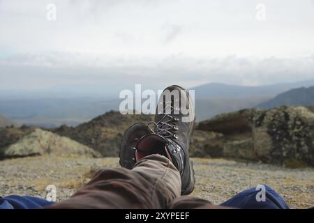 Gambe di un uomo che riposa in stivali per seguire le montagne sullo sfondo di montagne e valli con nuvole rumorose scarponi da trekking in montagna. Rif Foto Stock