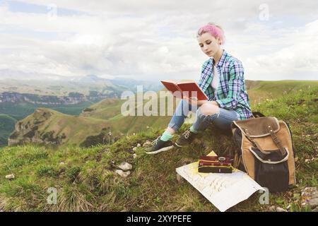 Una ragazza viaggiatrice siede tra le montagne sull'erba e legge un libro sullo sfondo di montagne epiche. Il concetto di lettura durante il riposo e le vacanze Foto Stock