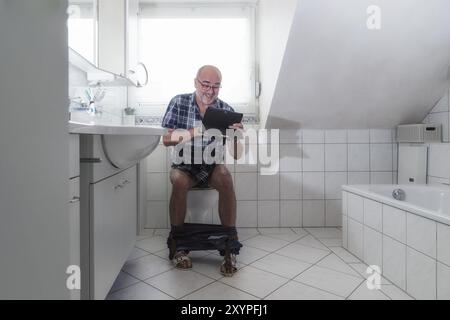 Un pensionato si siede a ridere in bagno e naviga su Internet su un tablet Foto Stock