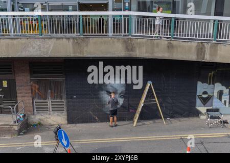 Southend on Sea, Regno Unito. 30 agosto 2024. Un artista di strada lavora su un murale monocromatico che raffigura il volto di un bambino su un muro di mattoni neri sotto un cavalcavia. La scena include coni stradali, una scala e un cartello stradale, con un passante che osserva dall'alto. Catturata a Southend-on-Sea, questa immagine mette in risalto la creatività urbana e il coinvolgimento del pubblico. Il primo giorno del City Jam si chiude con gli artisti che lavorano al loro lavoro creativo. Penelope Barritt/Alamy Live News Foto Stock