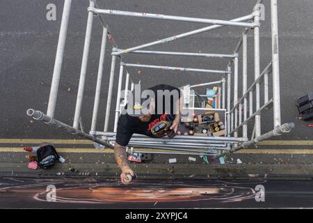 Southend on Sea, Regno Unito. 30 agosto 2024. Uno spruzzo di Street artist dipinge un realistico ritratto murale mentre si trova su un'impalcatura sopra una strada Southend-on-Sea. Scattata da un'angolazione elevata, l'immagine rivela gli strumenti, la tecnica e lo spazio di lavoro dell'artista, tra cui bombolette spray e attrezzatura di sicurezza. Le doppie linee gialle sottostanti suggeriscono l'ambientazione urbana e la scala dell'opera d'arte. Il primo giorno del City Jam si chiude con gli artisti che lavorano al loro lavoro creativo. Penelope Barritt/Alamy Live News Foto Stock