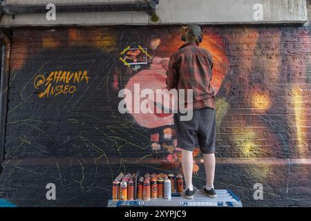 Southend on Sea, Regno Unito. 30 agosto 2024. Un artista di strada sta su una piattaforma mentre dipinge un vivace murale su una parete di mattoni. La grafica presenta un volto stilizzato con tonalità calde. Sono visibili diverse bombolette spray. Il primo giorno del City Jam si chiude con gli artisti che lavorano al loro lavoro creativo. Penelope Barritt/Alamy Live News Foto Stock