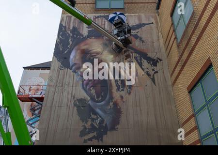 Southend on Sea, Regno Unito. 30 agosto 2024. Un grande murale che presenta un ritratto dettagliato viene dipinto sul lato di un edificio in mattoni a più piani. Un lavoratore in un sollevatore meccanico aggiunge tocchi di finitura, circondati da schizzi di vernice astratti. La scena cattura la trasformazione dello spazio urbano attraverso l'arte pubblica. Il primo giorno del City Jam si chiude con gli artisti che lavorano al loro lavoro creativo. Penelope Barritt/Alamy Live News Foto Stock