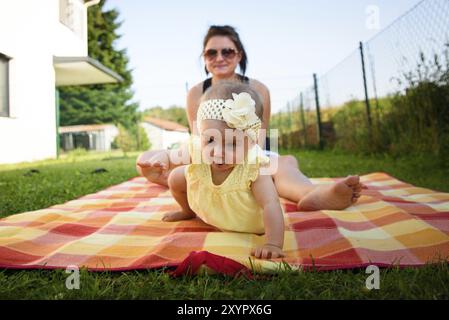 Ritratto giovane madre con cute baby girl in banda gialla e vestito su una coperta. Soleggiato giorno estivo all'aperto. Concetto di maternità Foto Stock