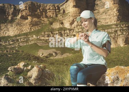 Ritratto di una giovane ragazza del fitness sorridente con un berretto e le cuffie che controllano il suo orologio intelligente seduto su una roccia all'aperto sullo sfondo di rocce Foto Stock