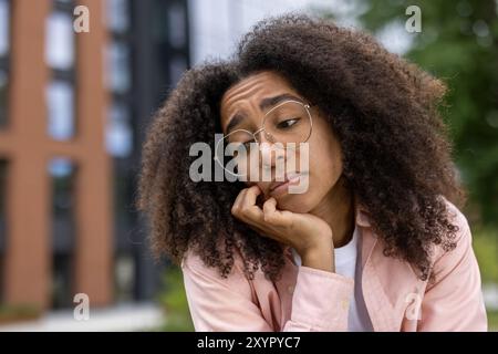 Donna afroamericana con capelli ricci e occhiali appare premurosa e contemplativa. Indossando un abbigliamento casual in città all'aperto, poggia il mento sulla mano, trasmettendo un pensiero profondo Foto Stock
