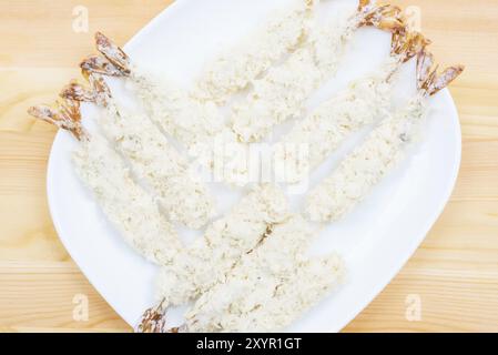 Primo piano di gamberi tempura crudi in un piatto impanato su un piatto bianco che si erge su un tavolo di legno. Semilavorati marini per la cucina casalinga Foto Stock