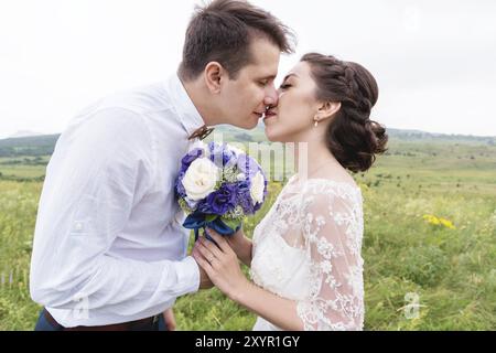 I giovani sposi novelli tengono insieme un bouquet di nozze e si baciano mentre sono iscritti in natura all'aperto Foto Stock