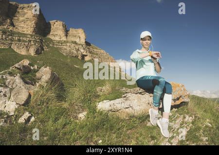 Ritratto di una giovane ragazza del fitness sorridente con un berretto e le cuffie che controllano il suo orologio intelligente seduto su una roccia all'aperto sullo sfondo di rocce Foto Stock