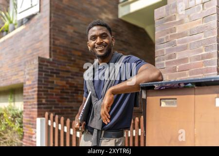 Giovane uomo d'affari africano appoggiato all'ingresso di una casa residenziale che porta con sé un computer portatile e sorride alla macchina fotografica Foto Stock