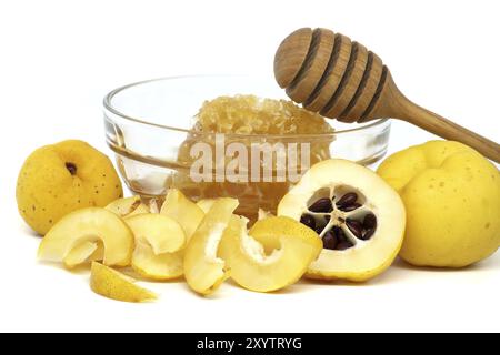 Ciotola di vetro piena di miele e cucchiaia di legno che riposa sul bordo circondato da vibranti frutti di cotogna, sia tagliati a fette che interi isolati su fondo bianco Foto Stock