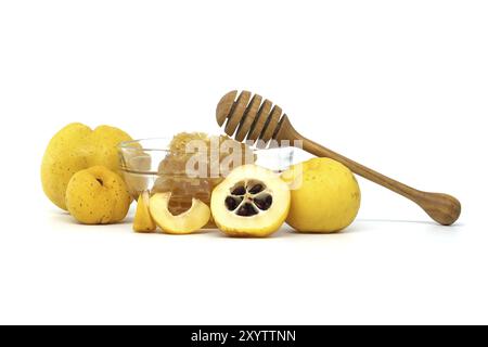 Ciotola di vetro piena di miele e cucchiaia di legno che riposa sul bordo circondato da vibranti frutti di cotogna, sia tagliati a fette che interi isolati su fondo bianco Foto Stock