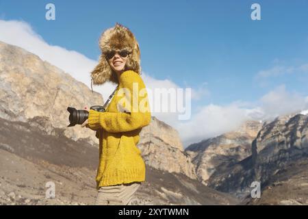 Una fotografa con occhiali da sole e un cappello di pelliccia grande e un maglione in maglia gialla si staglia sullo sfondo di alte rocce nella gola con una cameriera Foto Stock