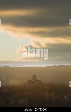 Paesaggio autunnale della parte del parco della città nella foschia pre-fumo dai toni caldi. Chiesa cattolica sullo sfondo di alti edifici Foto Stock