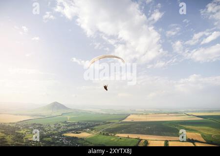 Il parapendio professionista in una tuta da cocco vola in alto sopra il terreno contro il cielo e i campi con le montagne Foto Stock