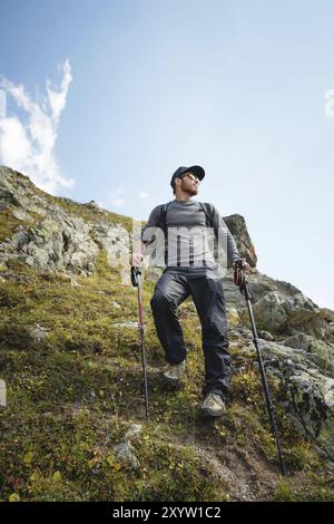 Un uomo barbuto con occhiali da sole e un berretto con uno zaino si staglia sulla cima di una roccia e guarda verso una valle rocciosa in alto tra le montagne. Il concetto di tour Foto Stock