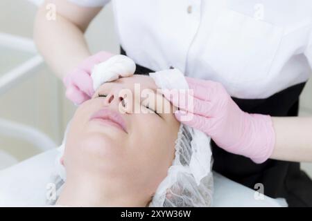 Un primo piano della procedura di pulizia nell'ufficio di cosmetologia. Le mani del cosmetologo in guanti rosa sono rimosse dal viso di un giovane Foto Stock